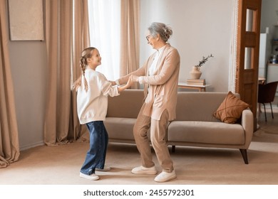 An older woman and a young girl are dancing in a cozy living room. The woman is wearing glasses and a floral dress while the girl is in casual clothes - Powered by Shutterstock