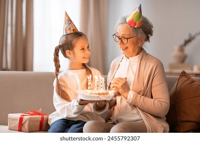 An older woman and a young girl are sitting on a couch together with a birthday cake placed in front of them. Both seem to be celebrating a special occasion together. - Powered by Shutterstock