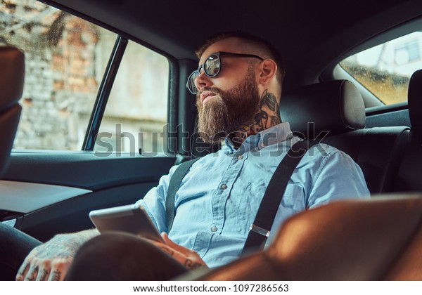 Old-fashioned tattooed hipster guy in a\
shirt with suspenders sitting in a luxury car on back\
seat.