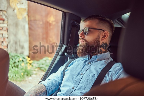 Old-fashioned tattooed hipster guy in a\
shirt with suspenders sitting in a luxury car on back\
seat.