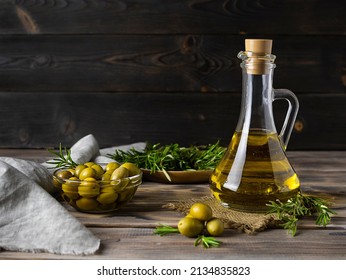 Olive Oil Of Golden Color In A Transparent Jug On A Canvas Napkin. Olives In A Glass Vase And Sprigs Of Rosemary In The Background. Dark Brown Wooden Background