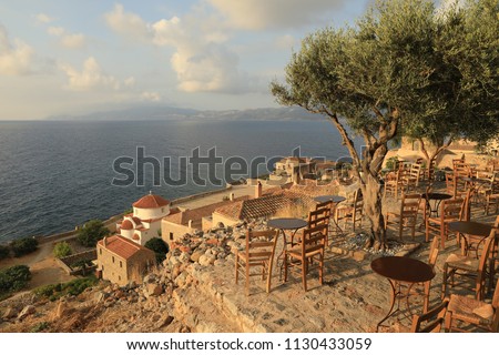 Olive tree surrounded by round iron tables and wooden chairs, typical traditional greek cafes, overlooking the roofs of houses and the sea in Monemvasia, Peloponnese, Greece, June 2018. Horizontal.