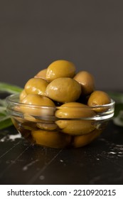 Olives In An Extra Virgin Olive Oil Placed In A Glass Bowl And Green Branches On A Dark Table With Copy Space. Vertical Shot, A Closeup.