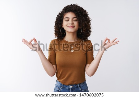 Om girl feels peace patience. Attractive carefree relaxed happy young woman curly shirt hairstyle close eyes smiling delighted meditating hands sideways nirvana lotus pose, breathing practice yoga