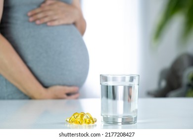 Omega 3 capsules and glass of water and pregnant woman at home interior. Healthy fatty acids nutritional supplement for prenatal support. Omega, DHA, vitamin D, fish oil for healthy pregnancy.