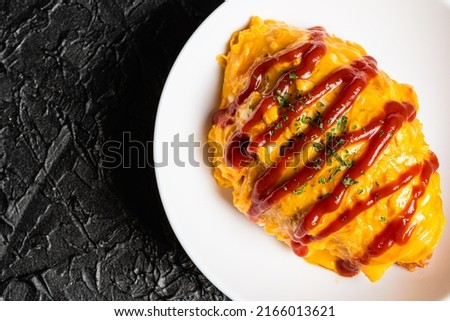 Omelette rice with tomato ketchup on a white plate against a black design board.