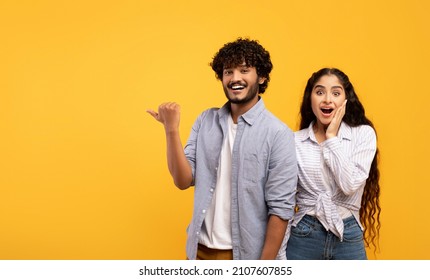 Omg, check this. Emotional indian guy pointing finger aside at free space over yellow background, surprised lady looking at camera, panorama. Couple demonstrating place for your text