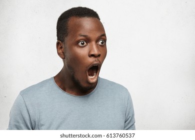 Omg. Half profile shot of pop-eyed funny young black man looking ahead of him with mouth wide opened and jaw dropped, his whole look expressing shock and full disbelief, standing at blank studio wall