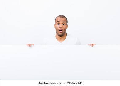 Omg! No Way! Advertising Concept. Confused Afro Man Is Standing Behind The Pure Blank Banner, Holding It, White Copyspace, In Casual Wear, Looking At The Camera, Shocked With Wide Open Eyes