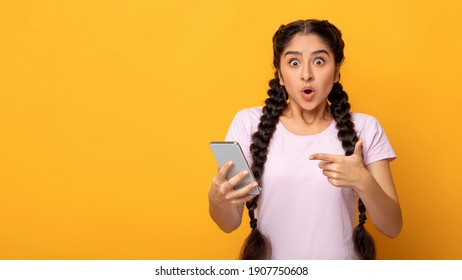 Omg. Portrait Of Emotional Shocked Indian Lady Holding And Using Mobile Phone, Pointing At Device. Surprised Woman Standing With Smartphone Isolated Over Yellow Studio Background, Banner, Copy Space