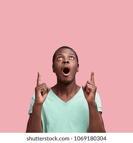 Omg and shock concept. Surprised African American male indicates at blank copy space upwads with fore fingers, isolated over pink wall, attracts your attention and asks to have look upwards.