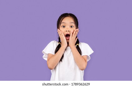 Omg. Shocked Asian Kid Girl Touching Face Looking At Camera In Amazement Standing Over Purple Background. Studio Shot