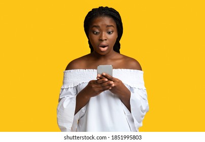 Omg. Surprised Black Woman Looking At Smartphone With Shocked Face Expression, Reading Message, Received Bad News, Got Unexpected Offer, Standing Over Yellow Studio Background, Copy Space