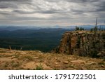 The ominous clouds reveal mountain silhouettes in the distance and the valley below the Mogollon Rim on FR 300 in Arizona. A large vertical cliff  at the edge of the Rim has several evergreens on top.