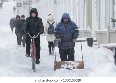 Omsk, Russia - January 11, 2018: The Cyclist Goes Down The Street In The Winter, A Row The Janitor Shovels Away Snow A Shovel