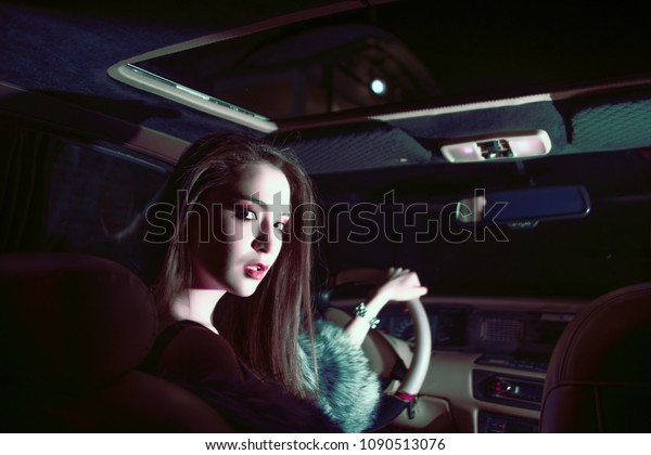 OMSK, RUSSIA -\
MARCH 14, 2017: Young woman drive and sitting in the seat of luxury\
car and looking at camera, against car window glass, travel and\
luxury concept. night\
scene