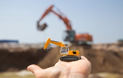 On The Arm Is A Small Orange Toy Excavator. Behind Him, Out Of Focus, Is A Real Working Excavator In A Quarry. A Bright Sunny Day. Operation Of Construction Equipment.