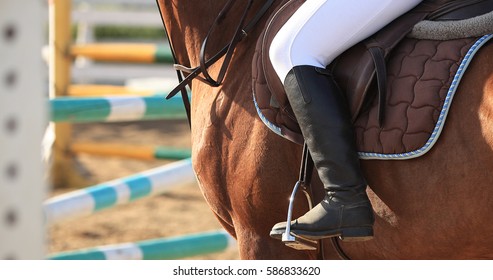 On back of a horse saddle, stirrup and leg rider Background with equipment. Obstacles, barriers to jumping. Riding on a Horse Equestrian Sport competitions showjumping. Sunny day, side view, close up.