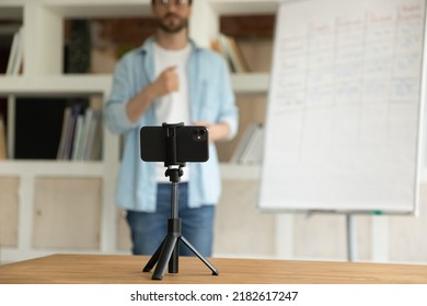 On Background Man Record Webinar On Cell Phone. Tutor Business Coach Stands Near Flip Chart Make Speech On Camera Filming Educational Video For Internet Audience. Vlogging, Tutoring, Education Concept