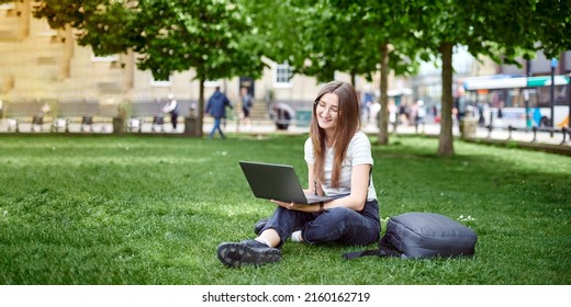 On the banner, a young girl works with a laptop in the fresh air in the park, sitting on the lawn. The concept of remote work. Work as a freelancer. The girl takes courses on a laptop and smiles. - Shutterstock ID 2160162719