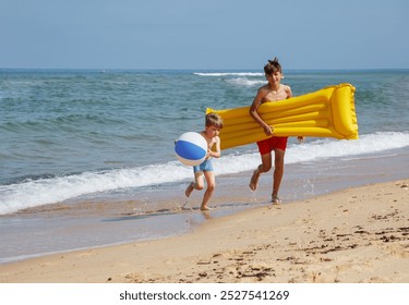 On the beach, two children dressed in swimwear outfits run toward the camera, holding various inflatable toys - Powered by Shutterstock