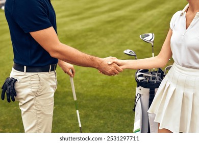 On a beautiful golf course, a man and woman shake hands, showcasing collaboration and sportsmanship essential to golf. Golf clubs and bags in the background enhance the atmosphere of this sport - Powered by Shutterstock