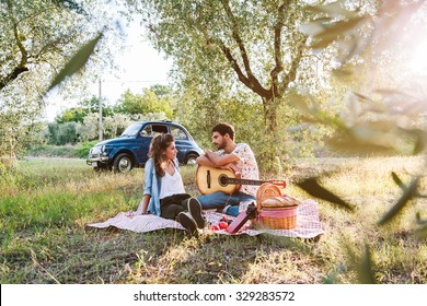 On a beautiful sunny day, a couple of young lovers, makes picnic on grass among olive groves in Tuscany, Italy. Man leans on guitar while talking with his girlfriend. Behind them a blue vintage car - Powered by Shutterstock