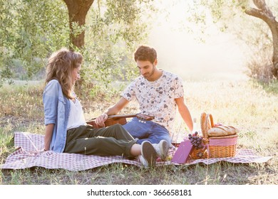 On a beautiful sunny day, a couple of young lovers, makes picnic on grass among olive groves in Tuscany, Italy. Man leans on guitar while talking with his girlfriend - Powered by Shutterstock