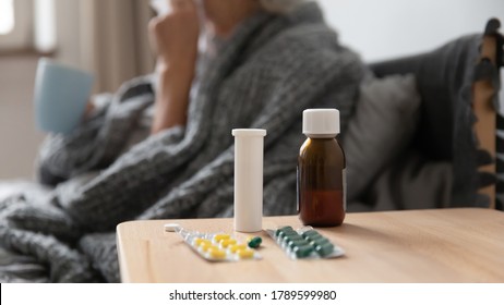 On bedside table blister pack with oral capsules syrup bottle close up, on background sick woman hold mug take antipyretic meds drink hot tea. Infected with grippe, corona virus being treated concept - Shutterstock ID 1789599980
