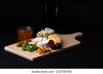 on a black background, a photograph was taken of a cheese platter laid on a wooden board, along with snacks - olives, nuts, grapes and basil leaves, next to it is a jar of natural bee honey
