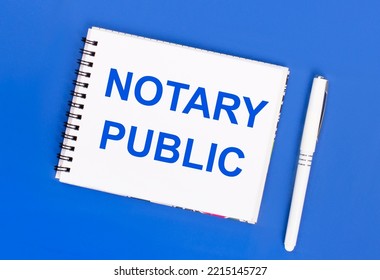 On A Blue Background, A White Ballpoint Pen And A White Notepad With The Text NOTARY PUBLIC. View From Above