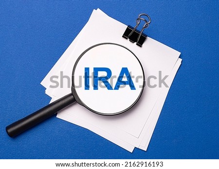 On a blue background, white sheets under a black paper clip and on them a magnifying glass with the text IRA Individual Retirement Account