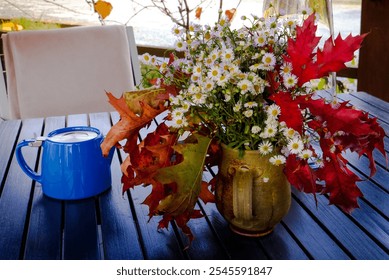 On a blue table, a large bouquet of white wildflowers sits in a ceramic vase, surrounded by vibrant, large leaves. Next to the bouquet stands a shiny blue pitcher. - Powered by Shutterstock