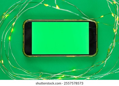 On a bright green background, a phone lies horizontally with a green chroma key screen surrounded by a garland that glows yellow. High quality photo