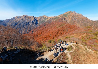 On a brisk autumn day, tourists gather at a viewpoint on a mountaintop to enjoy the view of Mount Tanigawa (谷川岳 Tanigawadake) under blue clear sky with fall colors on the mountainside, in Gunma, Japan