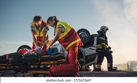 On the Car Crash Traffic Accident Scene: Paramedics Saving Life of a Female Victim who is Lying on Stretchers. They Apply Oxygen Mask, Do Cardiopulmonary Resuscitation / CPR and Perform First Aid