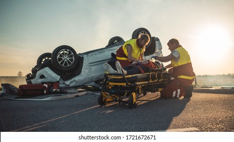 On the Car Crash Traffic Accident Scene: Paramedics Saving Life of a Female Victim who is Lying on Stretchers. They Listen To a Heartbeat, Apply Oxygen Mask and Give First Aid. Background Firefighters