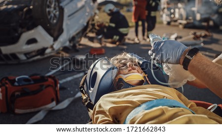 On the Car Crash Traffic Accident Scene: Paramedics Saving Life of a Traffic Accident Victim who is Lying on Stretchers. They Listen To a Heartbeat, Apply Oxygen Mask and Give First Aid Help