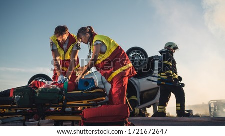 On the Car Crash Traffic Accident Scene: Paramedics Saving Life of a Female Victim who is Lying on Stretchers. They Apply Oxygen Mask, Do Cardiopulmonary Resuscitation / CPR and Perform First Aid