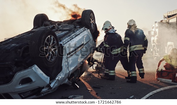On the Car Crash Traffic Accident: Paramedics and\
Firefighters Rescue Injured Trapped Victims. Medics give First Aid\
to Female on Stretchers. Firemen Use Hydraulic Cutters Spreader to\
Open Vehicle