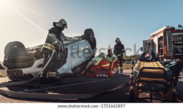 On the Car Crash Traffic Accident Scene: Team of\
Paramedics and Firefighters Rescue Injured People Trapped in\
Rollover Vehicle. Professionals Extricate Victims, give First Aid,\
Extinguish Fire