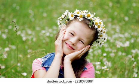 On A Chamomile Lawn, A Sweet Girl In A Wreath Of Daisies, Smiling, Pressing Her Hands To Her Cheeks. High Quality Photo