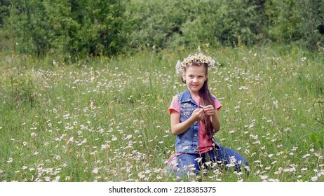 On A Chamomile Lawn, A Sweet Girl In A Wreath Of Chamomiles, Looks At The Daisy, Cuts Off The Petals, Sniffs It, And Smiles. High Quality Photo