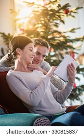 On Christmas morning, beautiful couple sitting in the living room, using a tablet, behind a decorated Christmas tree, the sun through the window give a cozy atmosphere