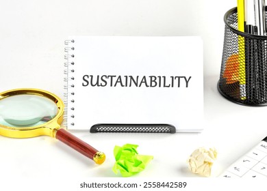 On a clean desk, a spiral notebook highlights the word sustainability. Nearby, a magnifying glass, crumpled paper, and assorted office supplies emphasize an eco-friendly theme. - Powered by Shutterstock