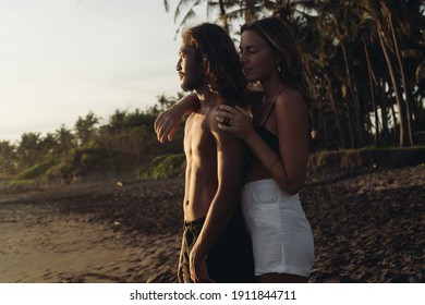 On The Coastal Black Sand Of Bali There Is A Beautiful Couple In Love. Woman Of European Appearance Hugs From The Back Of A Naked Torso Of A Man Of East Asian Appearance. High Quality Photo
