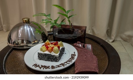 On The Coffee Table, On A White Plate, There Is A Cake Decorated With Cream And Fruit. There Is A Congratulatory Inscription On The Rim. Next To It Is A Metal Lid, Cutlery, A Plant In A Vase.