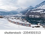 On a cold snowy winter day, Lucerne-Interlaken Express of Zentralbahn (ZB) travels thru Lungern village, with chalet houses on the lakeside and alpine mountains in background, in Obwalden, Switzerland