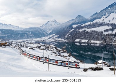 On a cold snowy winter day, Lucerne-Interlaken Express of Zentralbahn (ZB) travels thru Lungern village, with chalet houses on the lakeside and alpine mountains in background, in Obwalden, Switzerland