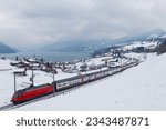 On a cold, snowy winter morning, a fast double-decker train travels along the shore of Lake Thun (Thunersee), with distant alpine mountains veiled by the clouds, in Spiez, Canton of Bern, Switzerland